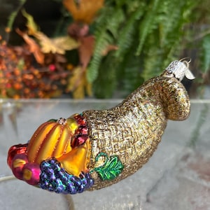 May include: A decorative cornucopia ornament with a gold basket weave design. The cornucopia is filled with colorful fruits and vegetables, including a pumpkin, grapes, and berries. A small silver leaf adorns the top. Perfect for fall or Thanksgiving decor.
