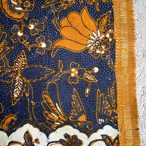 K&ouml;nnte beinhalten: Ein blauer und orangefarbener Stoff mit einem traditionellen indonesischen Batik-Muster. Das Muster zeigt V&ouml;gel, Blumen und Bl&auml;tter.