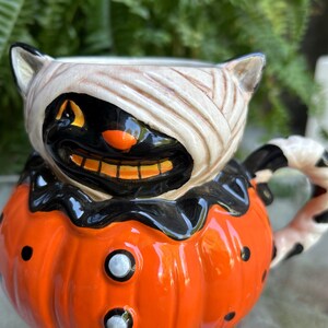 May include: A ceramic Halloween-themed mug. The mug is shaped like a pumpkin with a black cat face wrapped in bandages. The pumpkin is orange with black dots and white buttons. The handle has a black and white pattern.