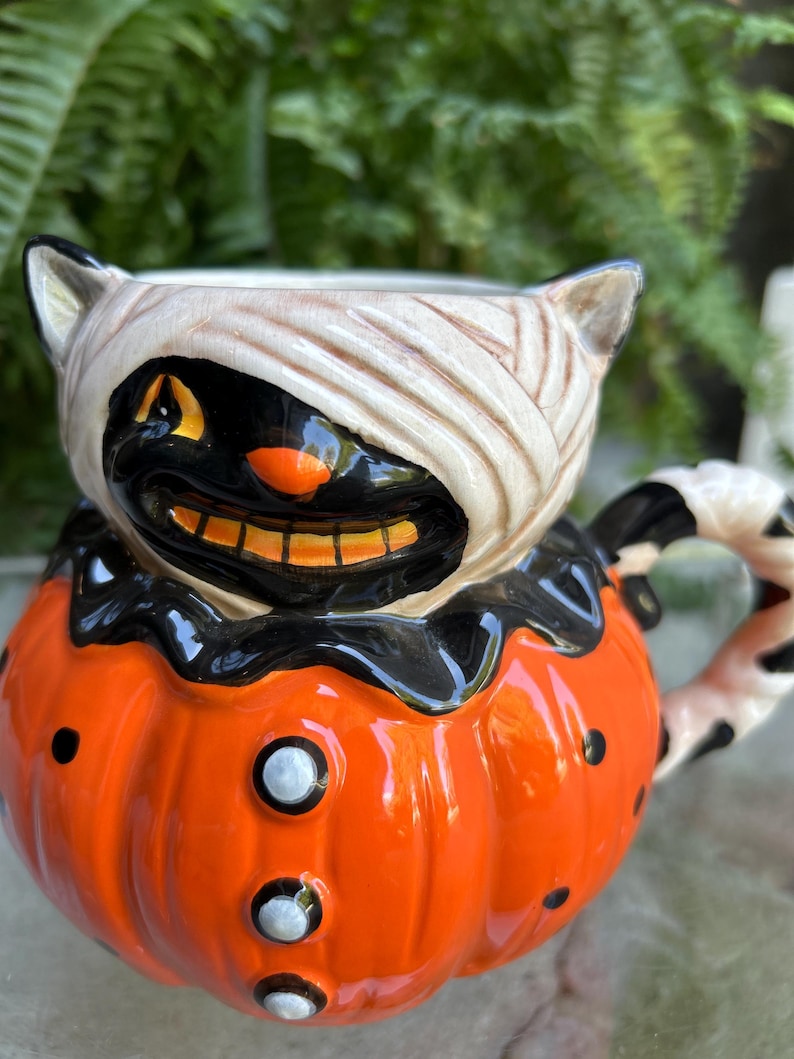 May include: A ceramic Halloween-themed mug. The mug is shaped like a pumpkin with a black cat face wrapped in bandages. The pumpkin is orange with black dots and white buttons. The handle has a black and white pattern.