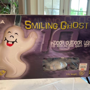 May include: A box for a set of 10 smiling ghost Christmas lights. The box is purple and features a large white ghost with a smiling face and a red mouth. The text on the box reads "Smiling Ghost 10 Indoor/Outdoor Use 10 Reflector Light Set 11 ft. length".