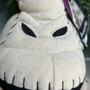 May include: Close-up of a plush Oogie Boogie doll. The doll is made of a beige burlap-like fabric with black stitched details and black eye cutouts. The mouth is a black, curved shape. A purple tag is visible.