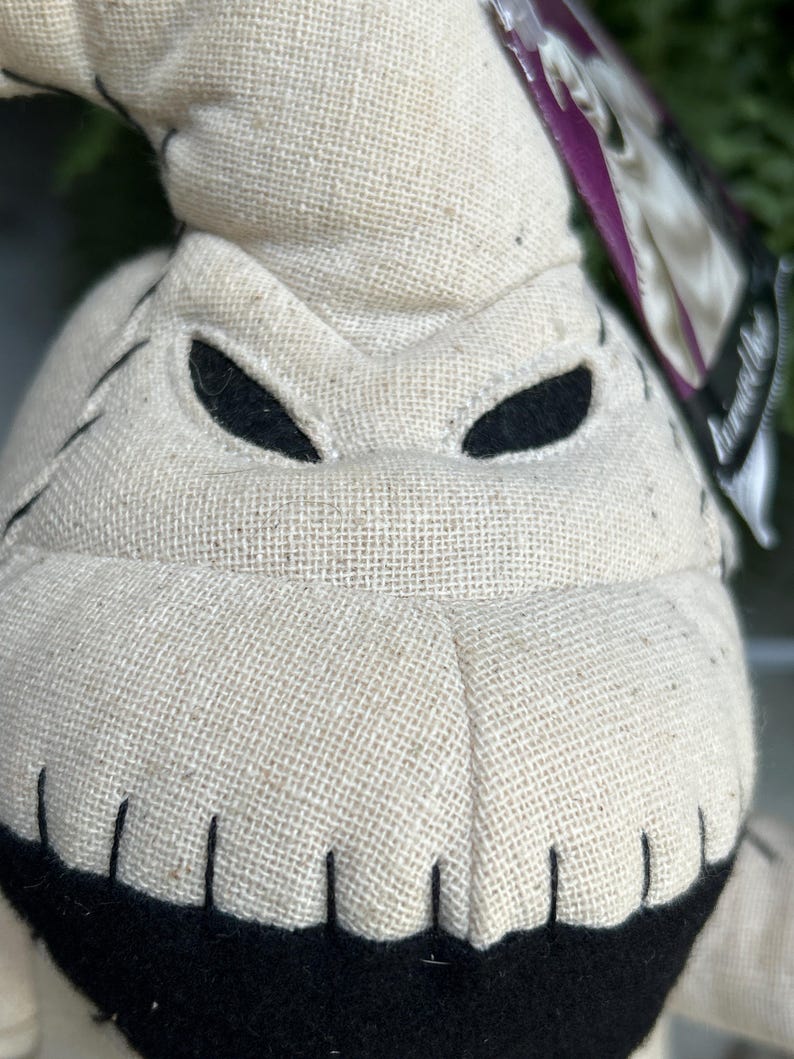 May include: Close-up of a plush Oogie Boogie doll. The doll is made of a beige burlap-like fabric with black stitched details and black eye cutouts. The mouth is a black, curved shape. A purple tag is visible.