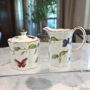 May include: A white porcelain sugar bowl with a lid and a creamer. Both pieces are decorated with red and blue butterflies, green leaves, and gold trim. The sugar bowl has a gold finial.