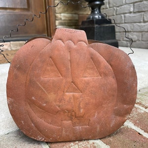May include: A rusty orange metal pumpkin with a carved jack-o'-lantern face and a wire handle. The pumpkin is sitting on a brick surface.