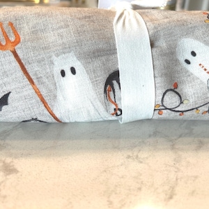 May include: Rolled-up Halloween-themed fabric with a white ribbon. The fabric features illustrations of ghosts, bats, and a pitchfork in orange and black on a light gray background. A string of lights is also visible.