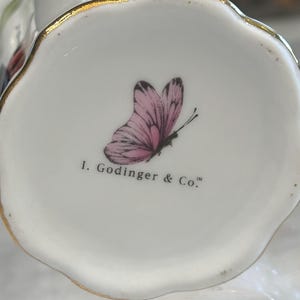 May include: Close-up of a white ceramic object with a scalloped edge and gold trim. A pink butterfly with black details is printed on the bottom, along with the text "I. Godinger & Co."