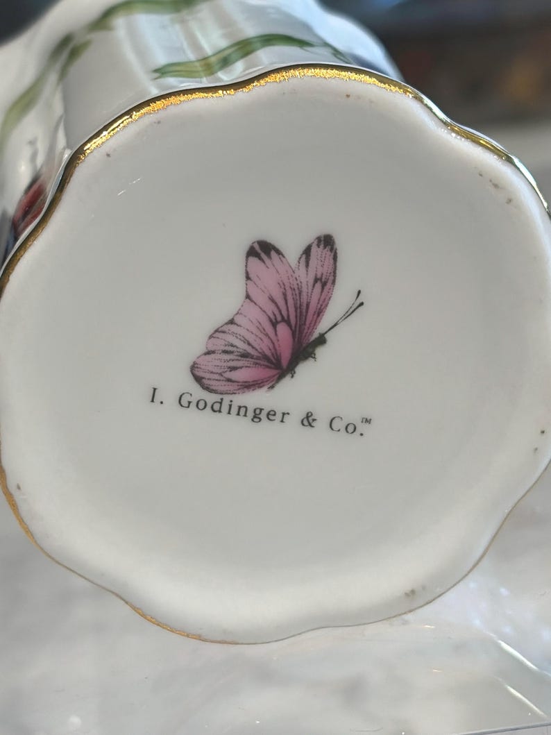 May include: Close-up of a white ceramic object with a scalloped edge and gold trim. A pink butterfly with black details is printed on the bottom, along with the text "I. Godinger & Co."