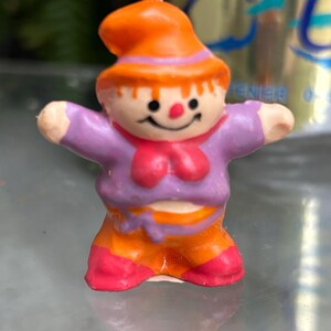 K&ouml;nnte beinhalten: Eine kleine, bunte Kerze in Form einer l&auml;chelnden Figur mit ausgestreckten Armen. Die Figur tr&auml;gt einen orangefarbenen Hut, ein lila Hemd, eine orangefarbene Hose und rosa Schuhe. Die Kerze hat einen Docht oben und ist vor einem verschwommenen Hintergrund platziert.