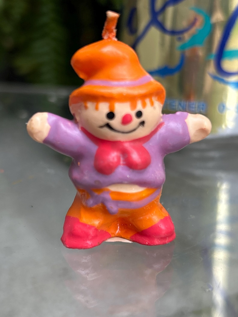 K&ouml;nnte beinhalten: Eine kleine, bunte Kerze in Form einer l&auml;chelnden Figur mit ausgestreckten Armen. Die Figur tr&auml;gt einen orangefarbenen Hut, ein lila Hemd, eine orangefarbene Hose und rosa Schuhe. Die Kerze hat einen Docht oben und ist vor einem verschwommenen Hintergrund platziert.