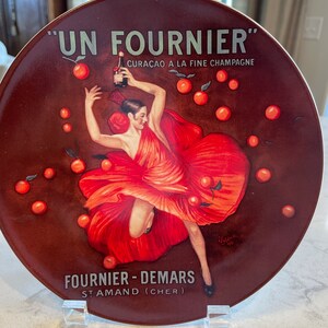 May include: A red and brown ceramic plate with a vintage illustration of a woman in a red dress holding a bottle of liqueur. The plate features the text "UN FOURNIER" CURAÇAO A LA FINE CHAMPAGNE, FOURNIER - DEMARS STAMAND (CHER).