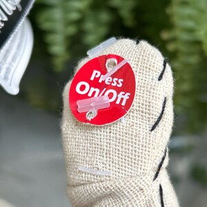 May include: Close-up of a stuffed fabric object with a red tag. The tag reads "Press On/Off" in white text. The object is beige with black stitching.