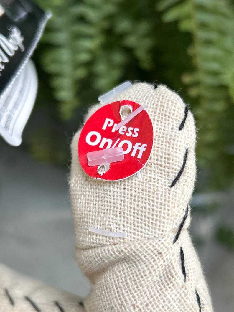 May include: Close-up of a stuffed fabric object with a red tag. The tag reads "Press On/Off" in white text. The object is beige with black stitching.