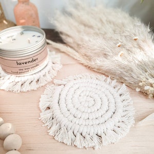 Può includere: Due sottobicchieri in crochet bianco con frange. I sottobicchieri sono rotondi e hanno un design a spirale. I sottobicchieri sono su una superficie in legno con una candela ed erba della pampa secca.