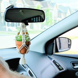 Könnte beinhalten: Eine kleine Topfpflanze hängt am Rückspiegel eines Autos. Die Pflanze befindet sich in einem Terrakotta-Topf und ist von einem Makramee-Hänger umgeben. Der Text "Bring your love of plants in your car!" ist im Vordergrund sichtbar.