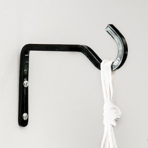 May include: Black metal plant hanger with a hook, mounted on a white wall. A white macrame plant hanger is hanging from the hook.