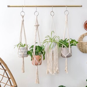 May include: Four macrame plant hangers with white cord and fringe hanging from a gold metal rod. Each hanger holds a potted plant. The plants are a variety of green foliage.