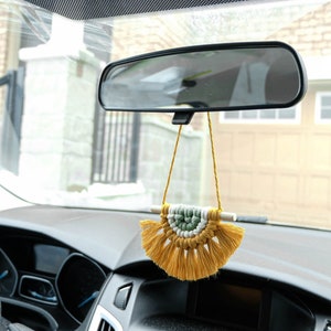 Op de afbeelding: Een gele en groene macramé autohanger die aan de achteruitkijkspiegel van een auto hangt.