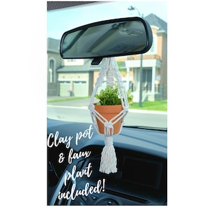 May include: White macrame plant hanger with a small terracotta pot and faux plant hanging from the rearview mirror of a car.  Clay pot & faux plant included!