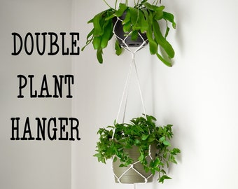 May include: A white macrame plant hanger with two hanging plants. The top plant is a green cactus-like plant in a black pot. The bottom plant is a green leafy plant in a brown pot. The text "DOUBLE PLANT HANGER" is written on the wall.