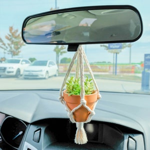 May include: A small potted plant hangs from the rearview mirror of a car. The plant is in a terracotta pot and is surrounded by a macrame hanger. The car's dashboard is visible in the foreground.