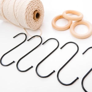 May include: A spool of natural twine, three wooden rings, and six black S-shaped hooks on a white surface.
