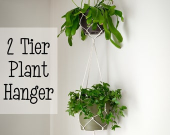 May include: A two-tier macrame plant hanger with two green plants in pots. The hanger is made of natural white cotton rope and is hanging from a hook on the ceiling. The text "2 Tier Plant Hanger" is written in black on a white background.