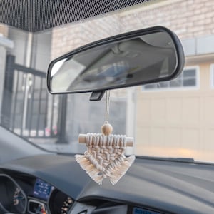 Może przedstawiać: Biały wisiorek do samochodu z makramą wiszący na lusterku wstecznym samochodu. Wisiorek wykonany jest z białego sznurka i ma drewnianą koralik na górze. Wisiorek ma kształt trójkąta z frędzlami na dole.