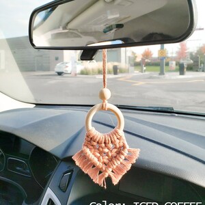 Może przedstawiać: Wisiorek do samochodu wykonany z makramu z drewnianym pierścieniem i frędzlami w jasnym brzoskwiniowym kolorze. Wisiorek jest zawieszony na lusterku wstecznym samochodu. Na dole obrazu widoczny jest tekst "color: ICED COFFEE".