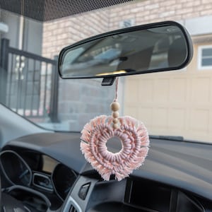 May include: A pink macrame heart-shaped car air freshener hanging from a car's rearview mirror. The charm has a circular centre and a fringed edge. Wooden beads are above the heart. The car's dashboard and steering wheel are visible.