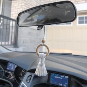 May include: A white tassel car charm with a wooden ring and a wooden bead. The charm is hanging from the rear view mirror of a car.