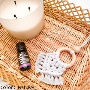 Puede incluir: Un ambientador de coche de macramé blanco con un anillo de madera y una borla. El ambientador está colgado de una cuenta de madera. El ambientador está sobre una superficie de mimbre con una botella de aceite esencial de lavanda.