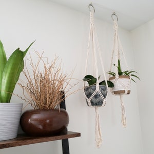 May include: Two macrame plant hangers with white cord and tassels, hanging from the ceiling. The hangers are holding two potted plants, one with green leaves and one with brown branches. The plants are in gray and white ceramic pots.