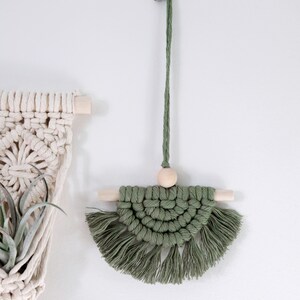 May include: A green macrame wall hanging with a wooden bead and wooden dowels. The hanging has a fringe around the bottom.