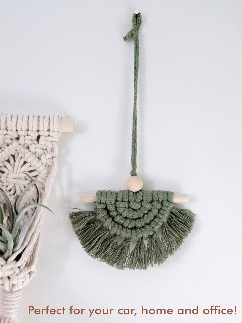 May include: A green macrame wall hanging with a wooden bead and wooden dowels. The hanging has a fringe around the bottom.