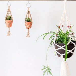 May include: Three macrame plant hangers with potted plants. The hangers are made of natural fibre rope and have wooden beads. The plants are green and leafy.