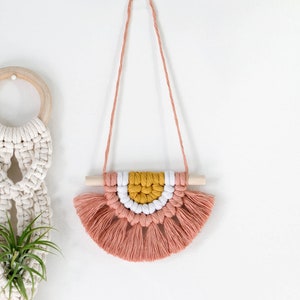 May include: A pink, white, and yellow macrame wall hanging with a wooden dowel and tassels. The hanging is attached to a white wall with a clear adhesive hook.