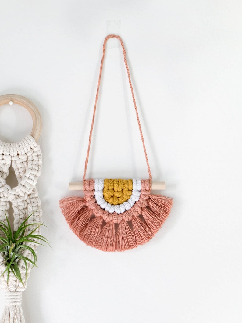 May include: A pink, white, and yellow macrame wall hanging with a wooden dowel and tassels. The hanging is attached to a white wall with a clear adhesive hook.
