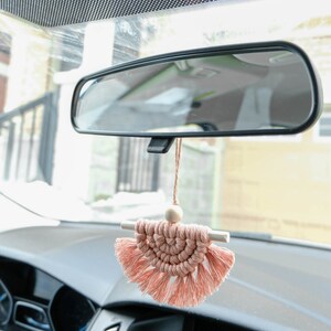 May include: A pink macrame car charm with a wooden bead and tassels hanging from the rearview mirror of a car.