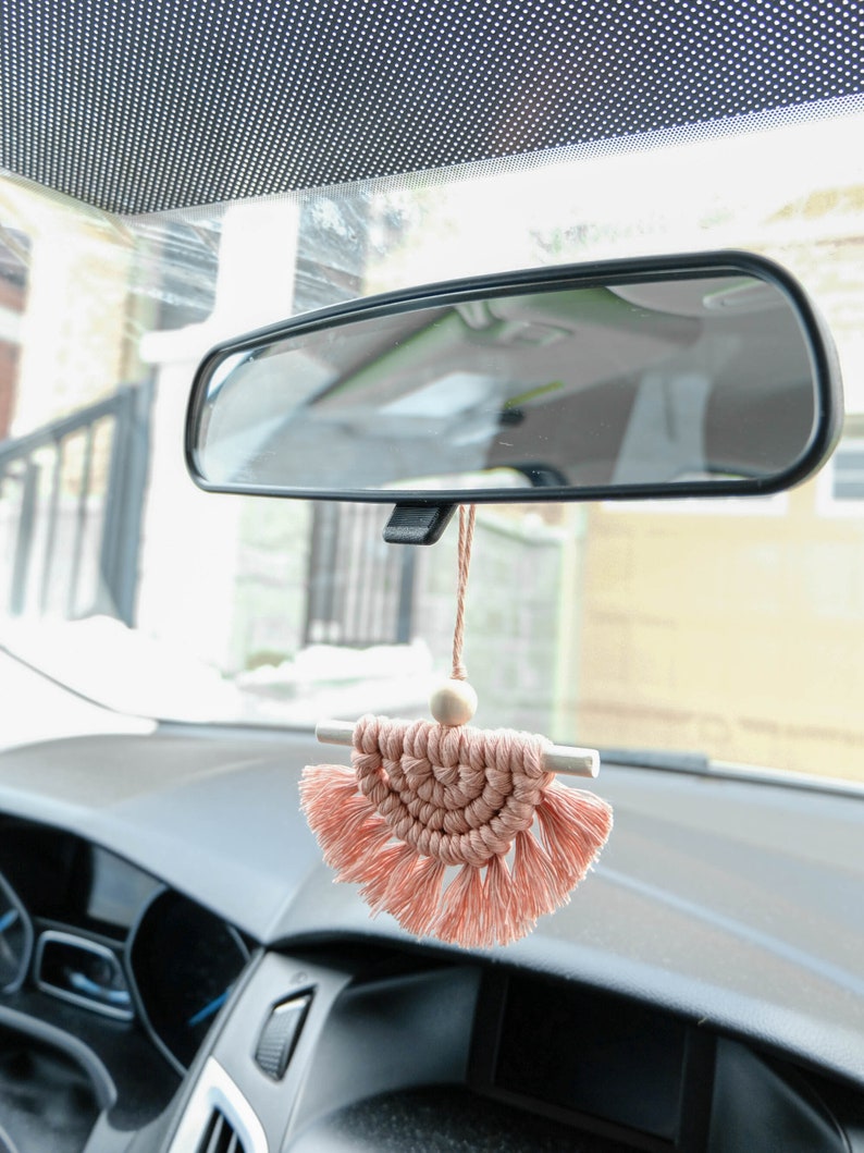May include: A pink macrame car charm with a wooden bead and tassels hanging from the rearview mirror of a car.
