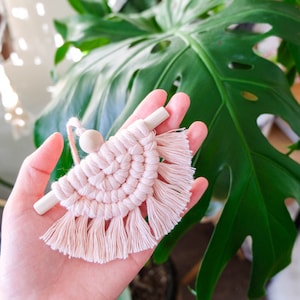 May include: A pink macrame wall hanging with a wooden dowel and a wooden bead. The wall hanging has a fringe around the bottom.