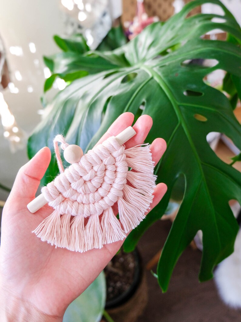 May include: A pink macrame wall hanging with a wooden dowel and a wooden bead. The wall hanging has a fringe around the bottom.