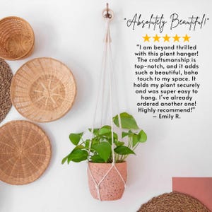 May include: A macrame plant hanger with a pink speckled pot and a green plant hanging from it. The hanger is made of natural cotton rope and has a simple, bohemian style. The background features a white wall with wicker baskets and mats.