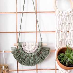 May include: A green and white macrame wall hanging with tassels, hanging on a copper wire grid. The wall hanging is in the shape of a half circle and has a wooden dowel at the top.
