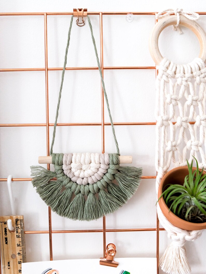 May include: A green and white macrame wall hanging with tassels, hanging on a copper wire grid. The wall hanging is in the shape of a half circle and has a wooden dowel at the top.