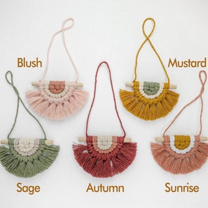 May include: Six macrame wall hangings with wooden dowels and fringe. The colors are blush, sage, autumn, sunrise, and mustard.