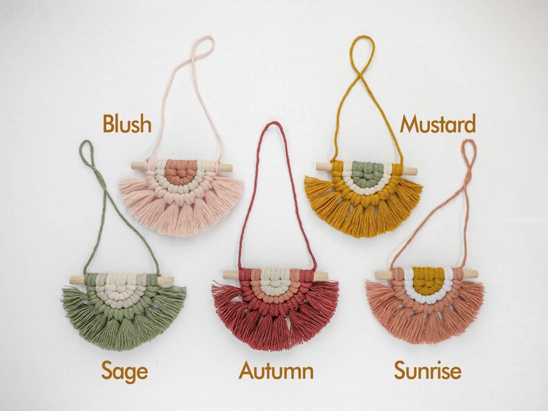 May include: Six macrame wall hangings with wooden dowels and fringe. The colors are blush, sage, autumn, sunrise, and mustard.