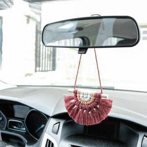 May include: A pink and white macrame car charm hanging from the rearview mirror of a car. The charm is in the shape of a half-circle with tassels.