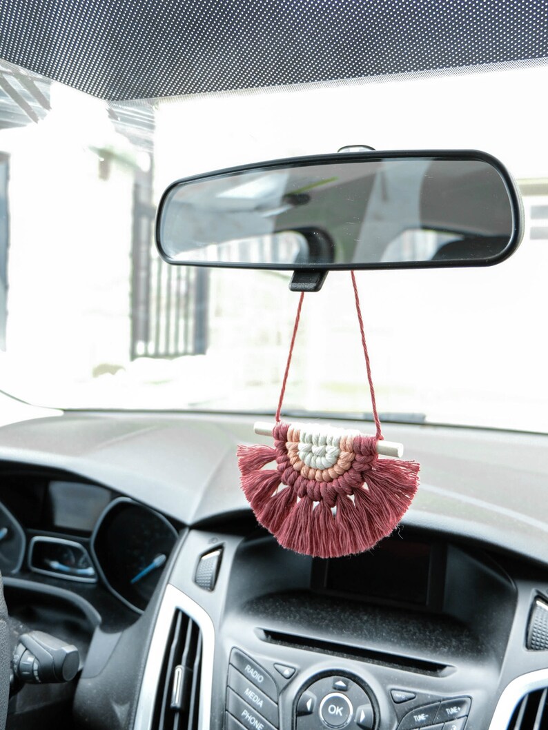 May include: A pink and white macrame car charm hanging from the rearview mirror of a car. The charm is in the shape of a half-circle with tassels.