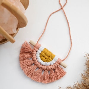 May include: A pink and yellow macrame wall hanging with a wooden dowel and fringe. The hanging is attached to a string.